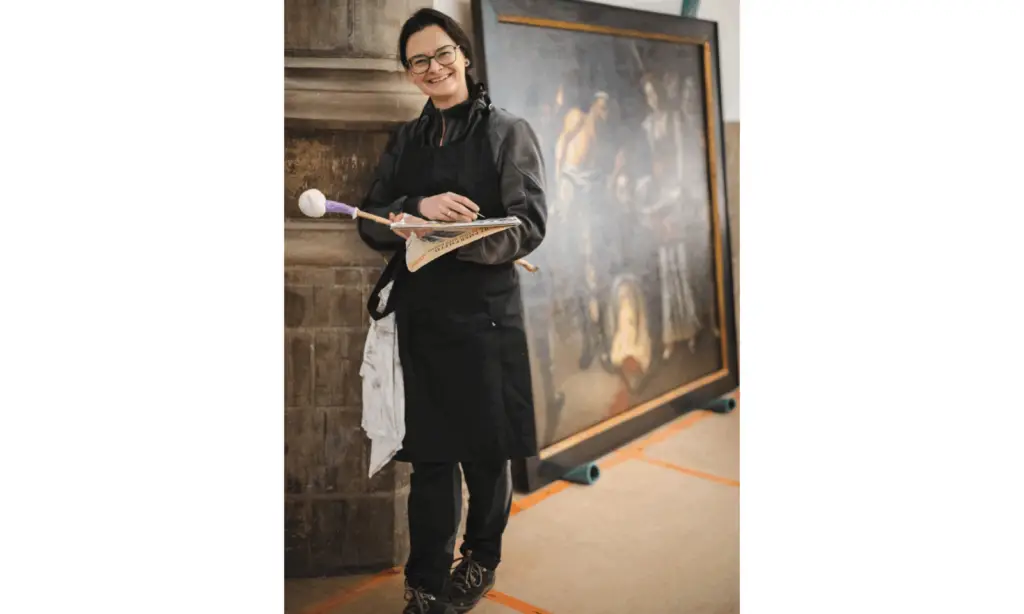 Restauratiespecialist met kwast in de hand werkt aan een historisch kunstwerk in een erfgoedproject, met een groot schilderij en monumentale stenen structuur op de achtergrond.