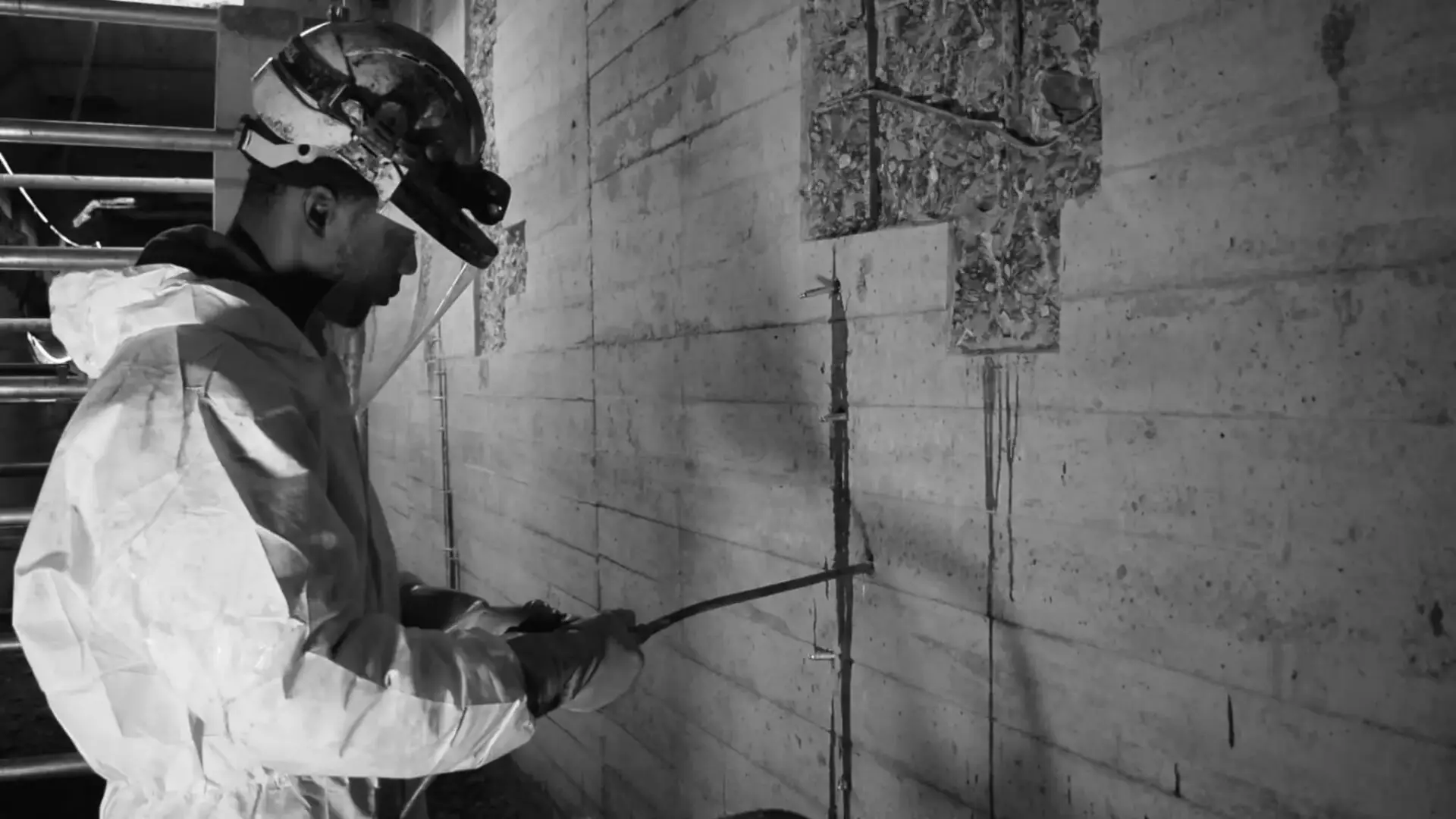 Vakman die injectiewerken uitvoert op een betonnen wand, met handgereedschap en beschermende werkkledij.