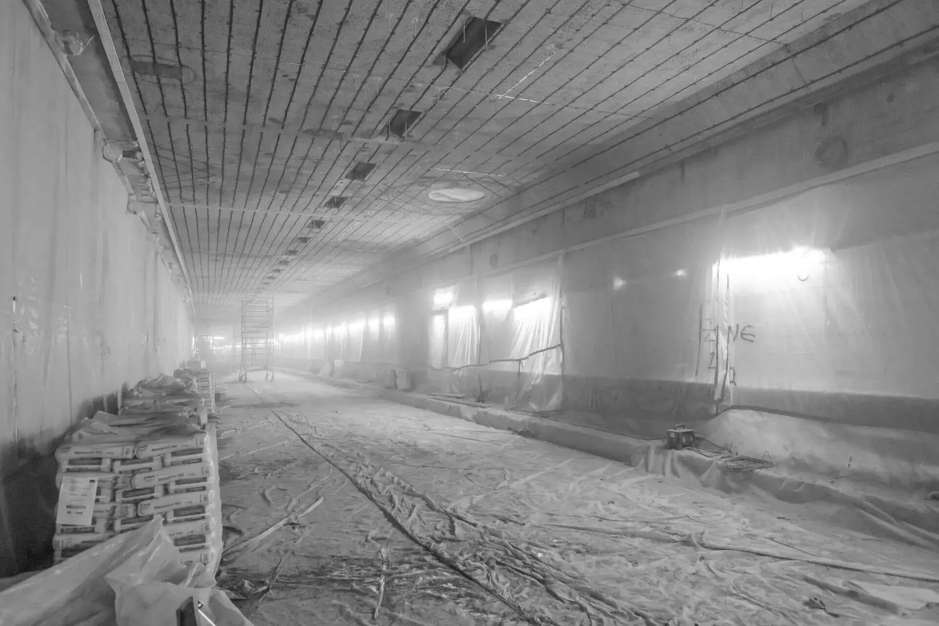 Grote tunnelruimte volledig afgeschermd met beschermfolie langs de wanden, met een plastic afdeklaag over de vloer, stapels bouwmaterialen en een steiger op afstand, gebruikt tijdens betonrenovatie met kathodische bescherming.