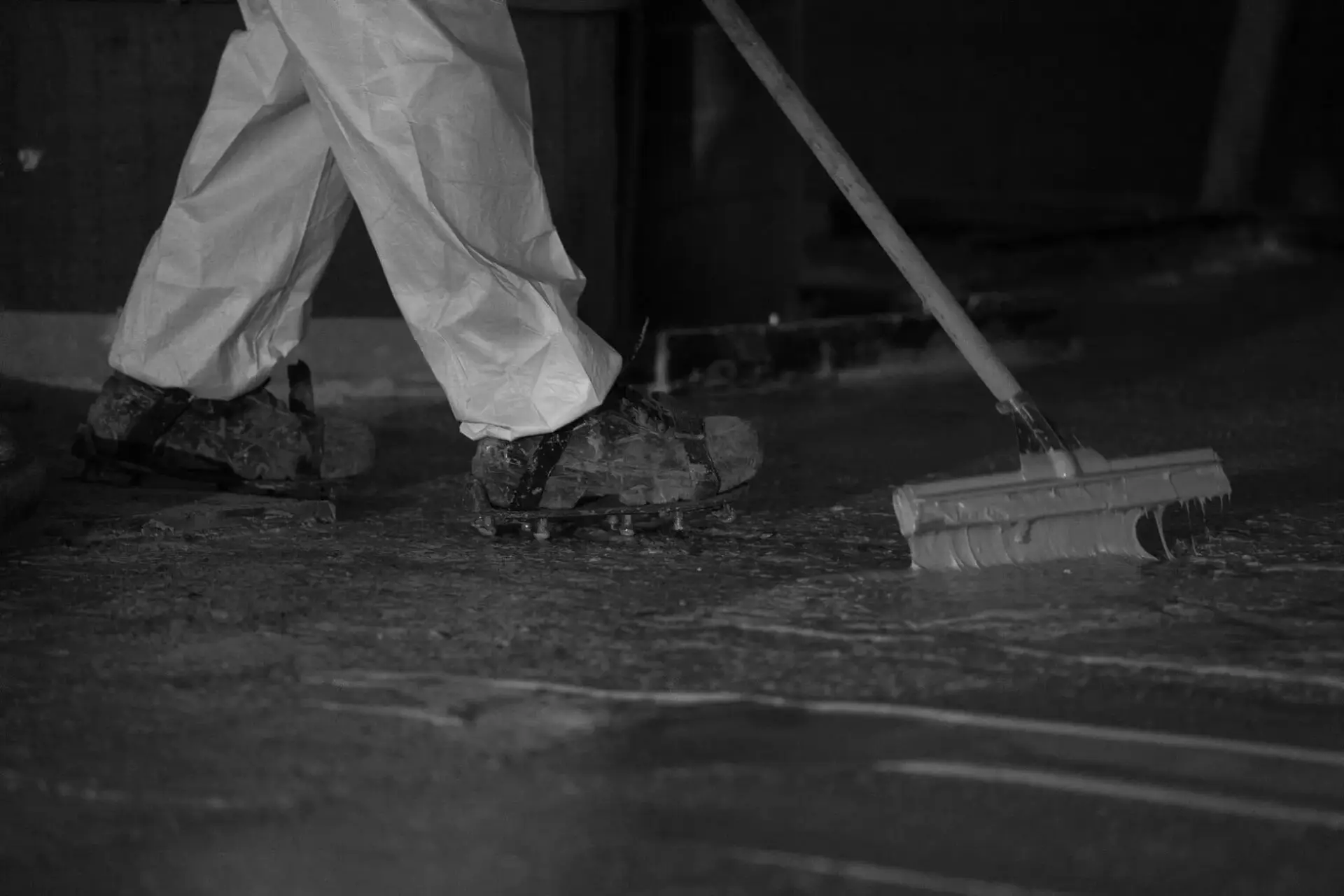 Detailopname van waterdichtingswerken waarbij een medewerker op spijkerzolen een vloeibare coating verdeelt over een betonvloer met een getande rakel, als onderdeel van renovatiewerken.